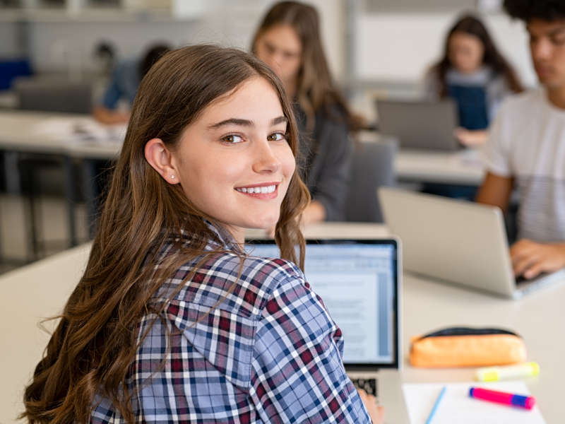 A smiling female student turns to look at the camera while working on a laptop in a classroom.